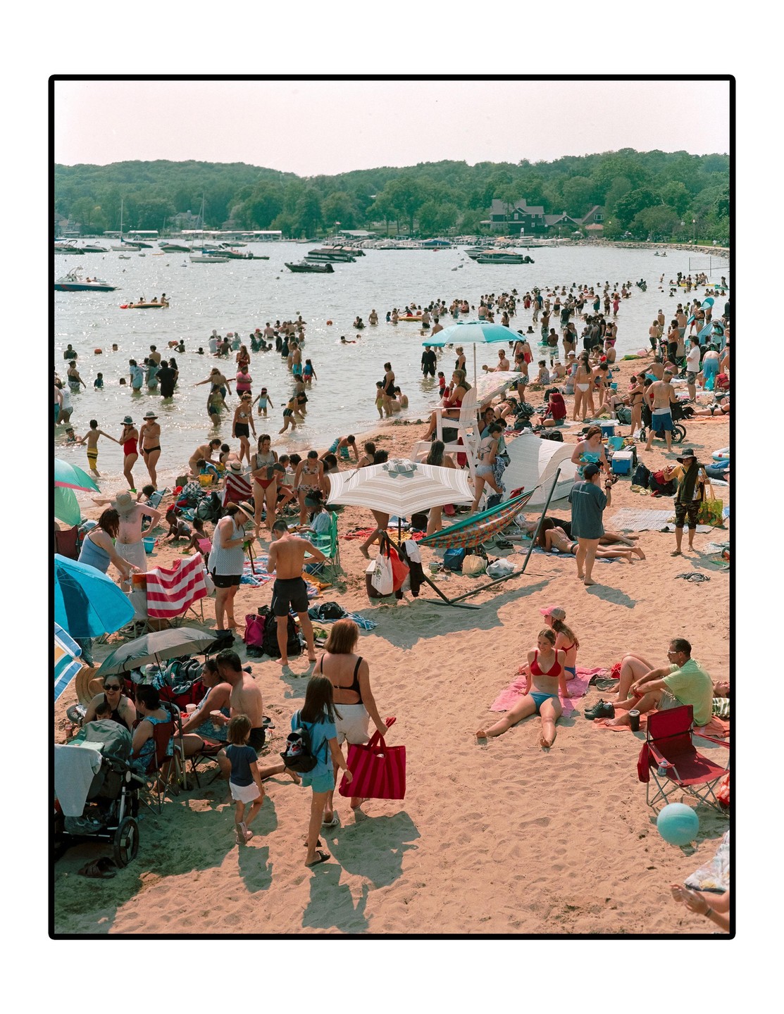 A crowded beach is filled with people swimming and relaxing on the sand, with boats visible in the water and trees on the distant shore.