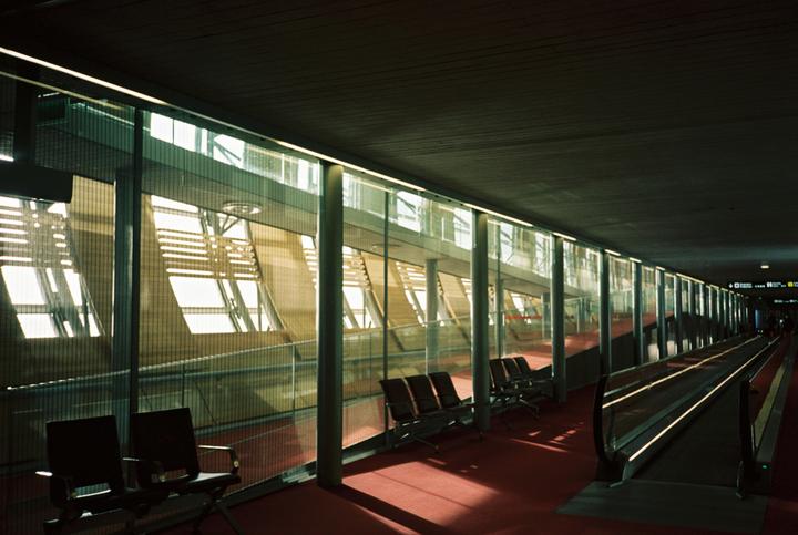 Sunlight streams through large windows onto a red carpeted floor in an airport terminal with rows of empty seats and a moving walkway.