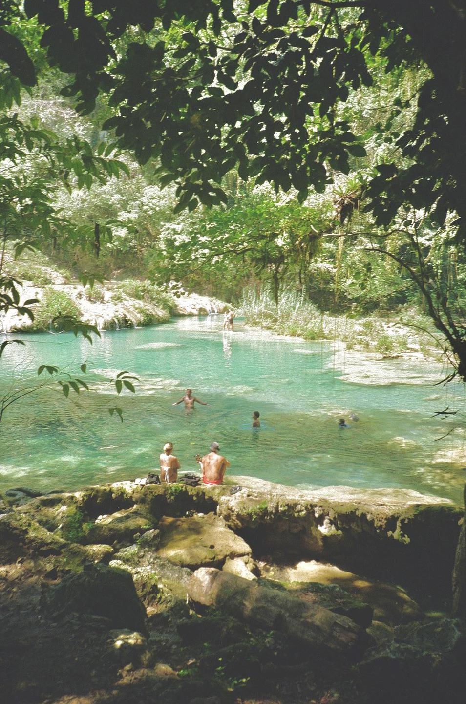 People swim and relax in a clear blue-green river surrounded by lush green foliage.