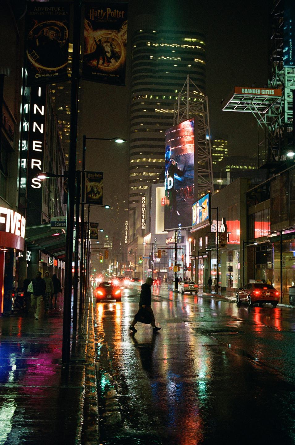 A person crosses a wet city street at night, reflecting the neon lights of surrounding buildings and cars.