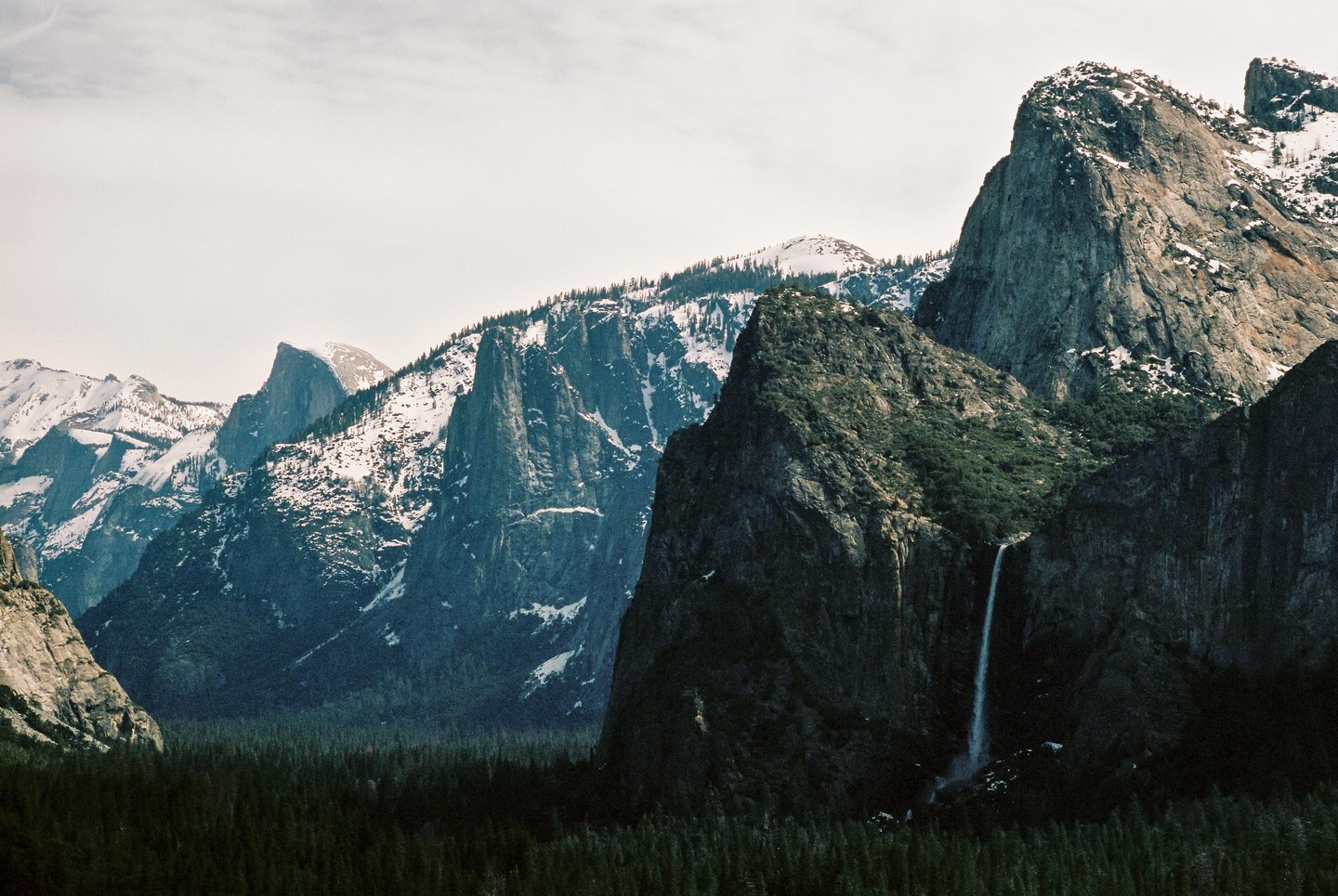 A waterfall cascades down a large mountain into a valley of evergreen trees, with snow-capped mountains in the background.