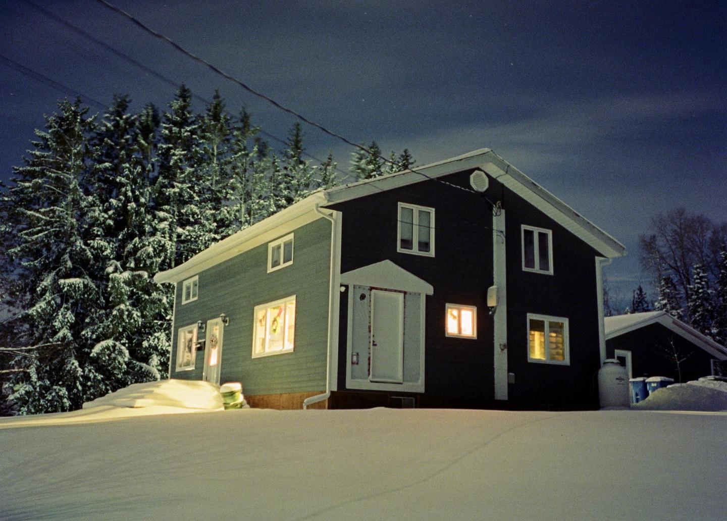 A house with illuminated windows stands in a snow-covered landscape at night, surrounded by snow-dusted evergreen trees.