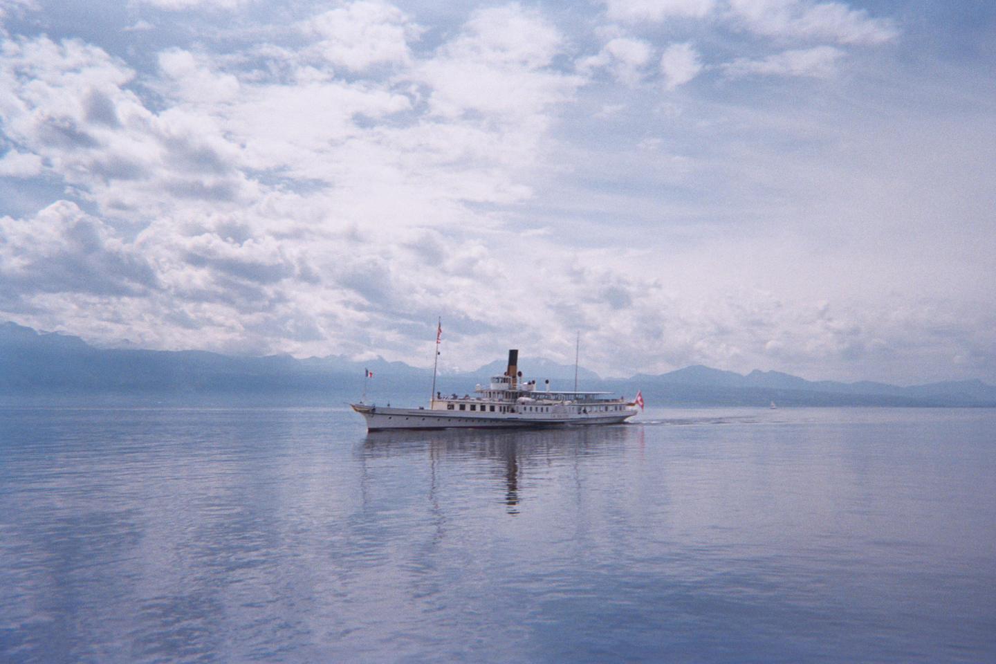 A white ferry boat with a black smokestack travels across a calm lake under a cloudy sky with mountains in the distance.