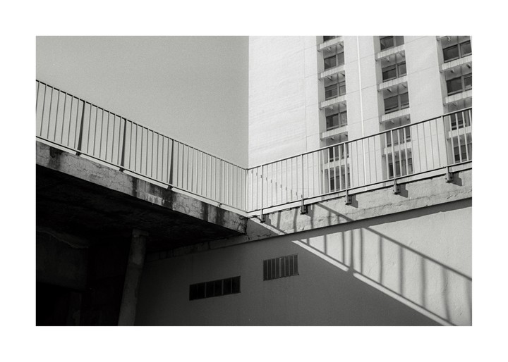 A concrete walkway with a metal railing casts a shadow on a building wall next to a tall building with many windows.
