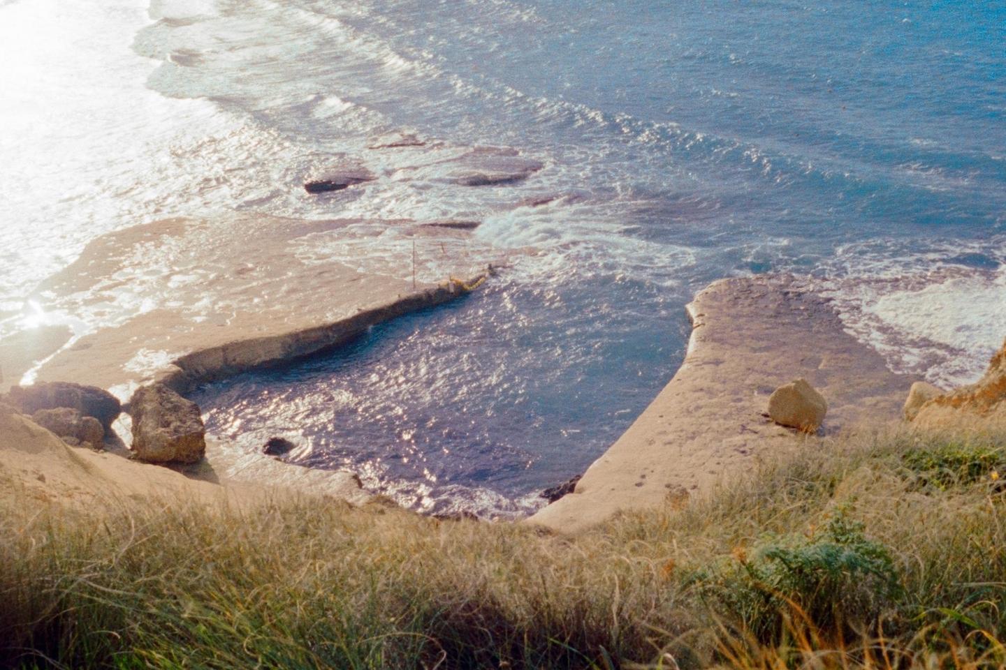 A natural pool carved into the rock is filled with ocean water, seen from a grassy cliff.