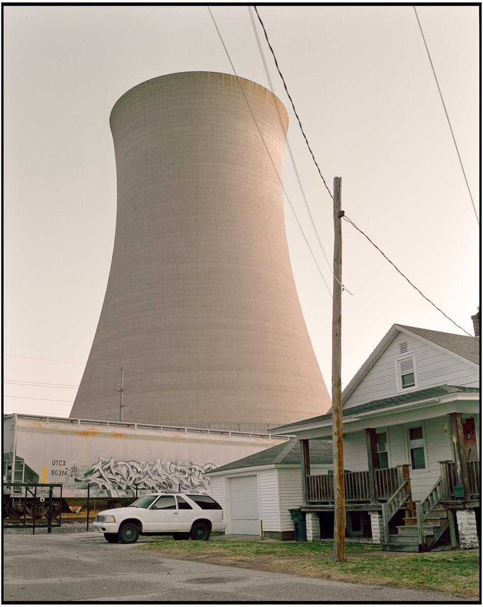 A large cooling tower looms over a residential street with a white house, a white car, and a train car with graffiti.