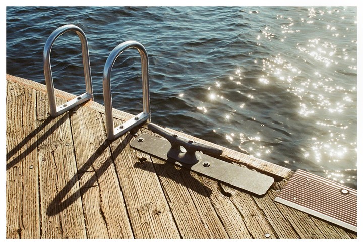 A wooden dock with a metal ladder and a cleat extends over sparkling water.