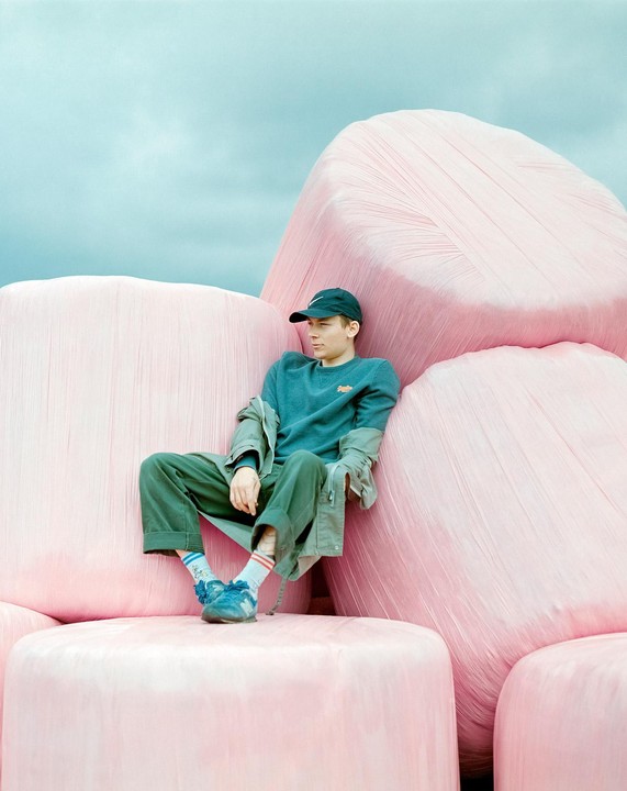 A man in a cap and sweatshirt sits on a stack of pink hay bales under a blue sky.