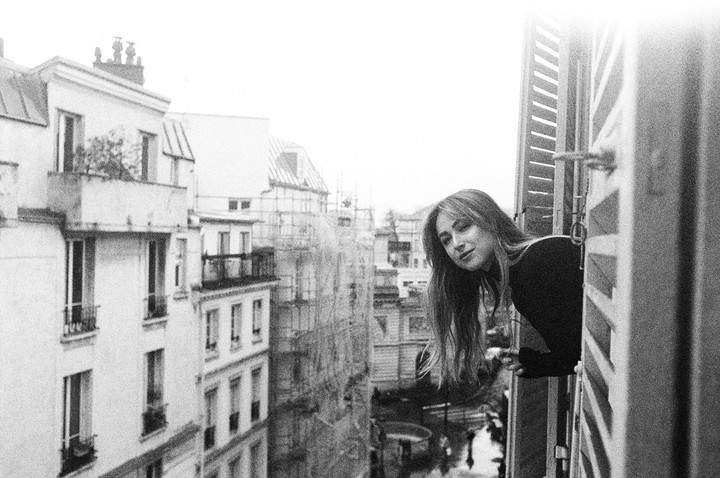 A woman leans out of a window with open shutters, looking towards the viewer, with Parisian buildings in the background.