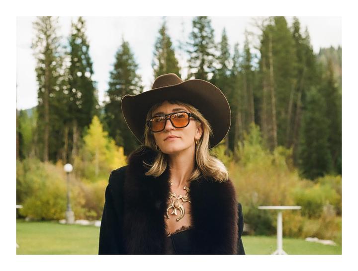 A woman wearing a cowboy hat and sunglasses stands in a grassy area with trees in the background.