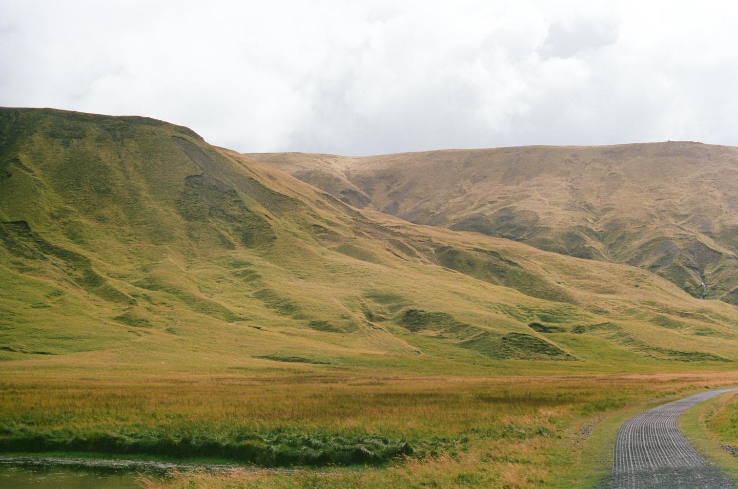 A gravel road curves through a grassy field with rolling hills under a cloudy sky.
