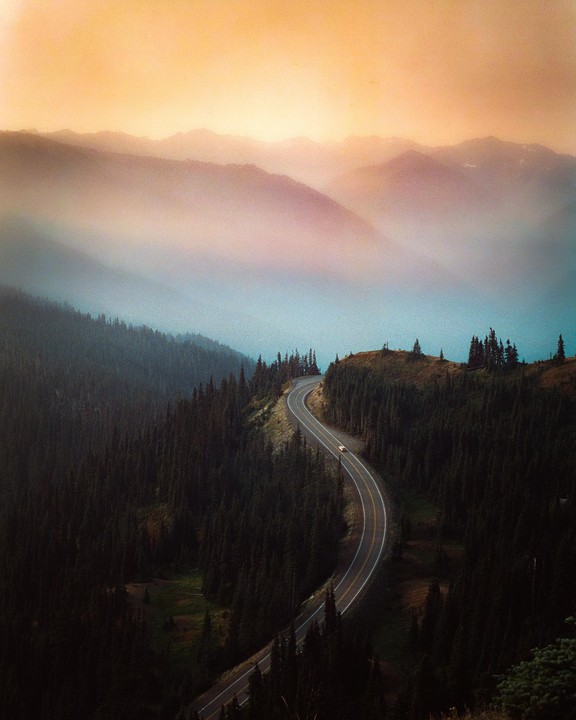 A winding road cuts through a dense forest with hazy mountains in the background.