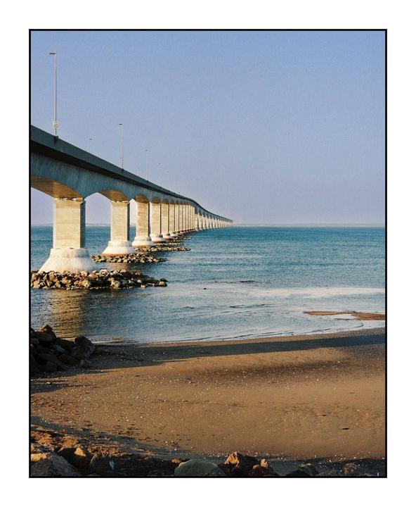 A long bridge extends over the water next to a sandy beach with rocks.