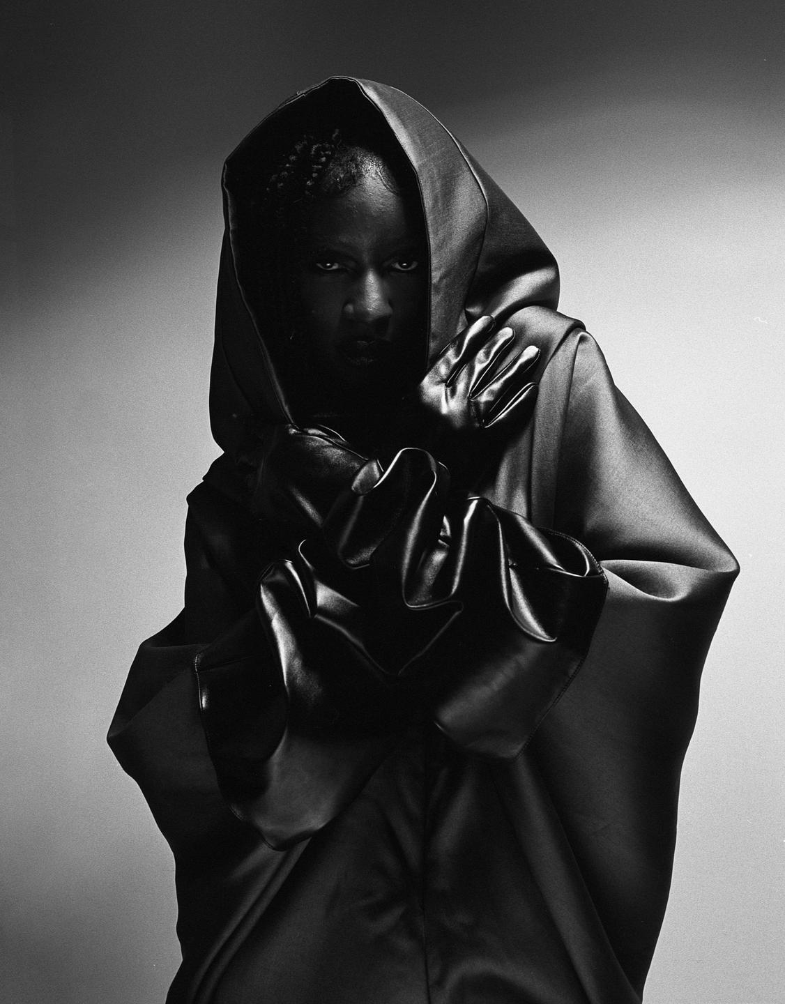 A person in a dark hooded robe and gloves looks directly at the viewer.