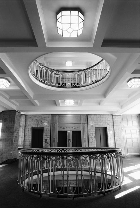 A black and white photo shows the interior of a building with a circular balcony and multiple elevators.