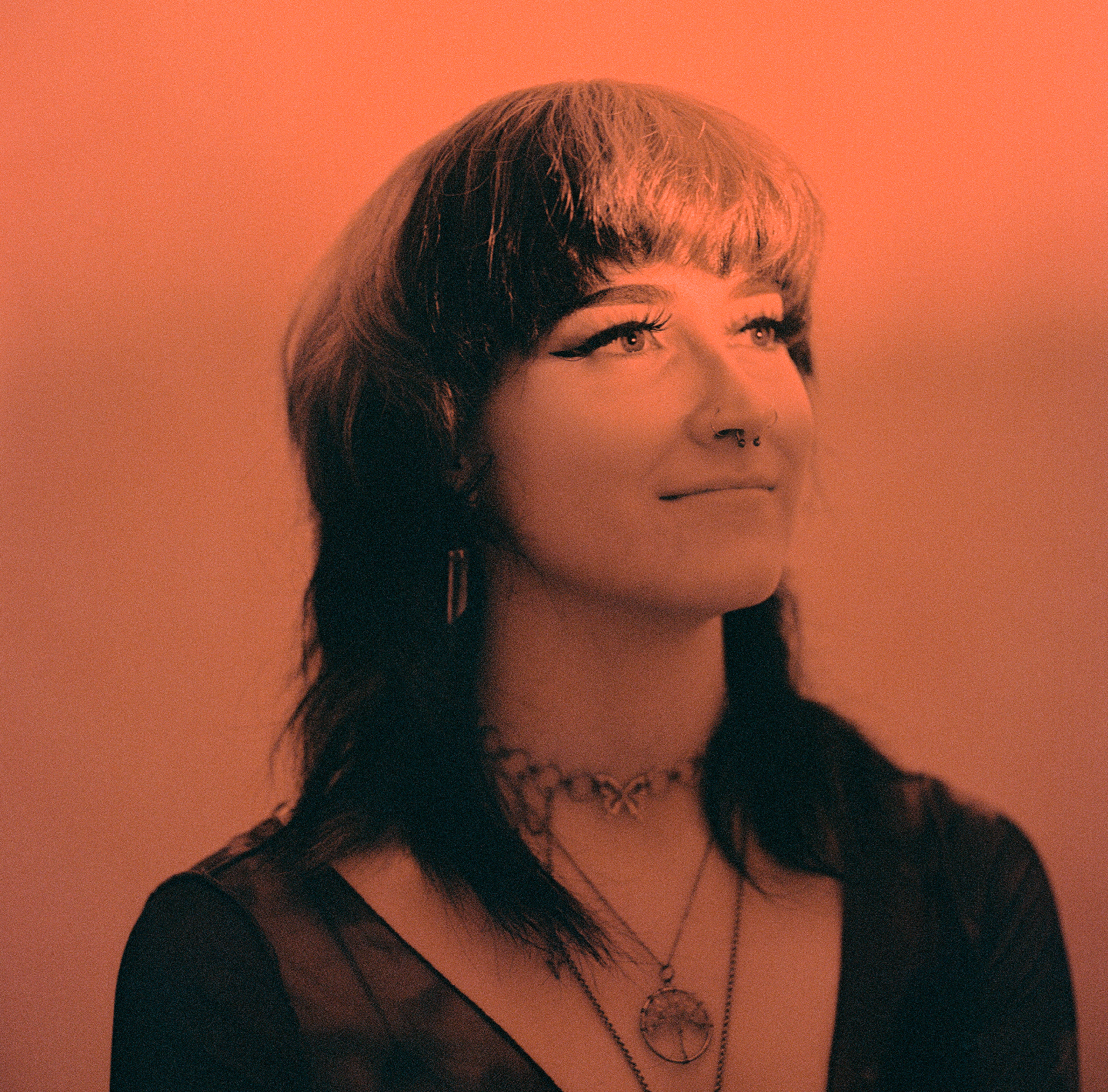A woman with a nose ring and necklaces looks up and to the right.