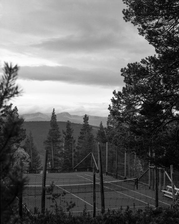 A tennis court is visible through trees with mountains in the background under a cloudy sky.