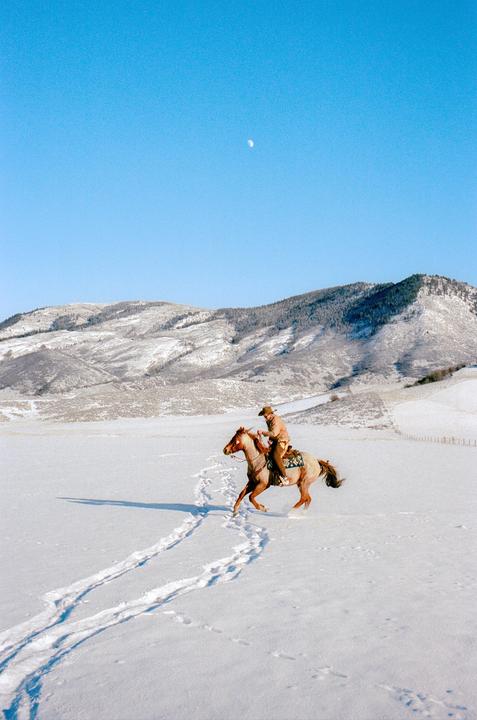 A man on horseback rides through a snow-covered field with mountains in the background under a clear blue sky with a visible moon.