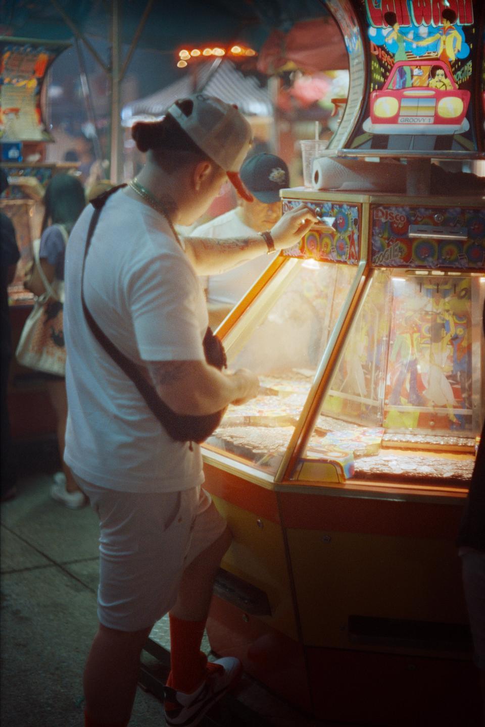 A man in a white shirt and baseball cap plays a coin pusher arcade game at night.