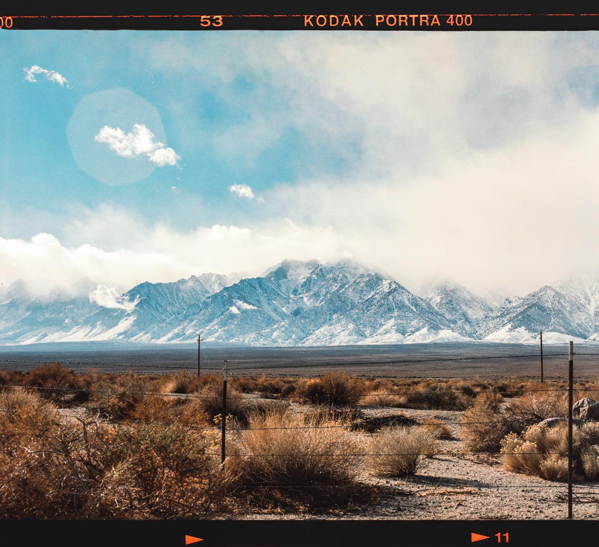 A vast desert landscape with sparse vegetation in the foreground and snow-capped mountains under a cloudy sky in the background.