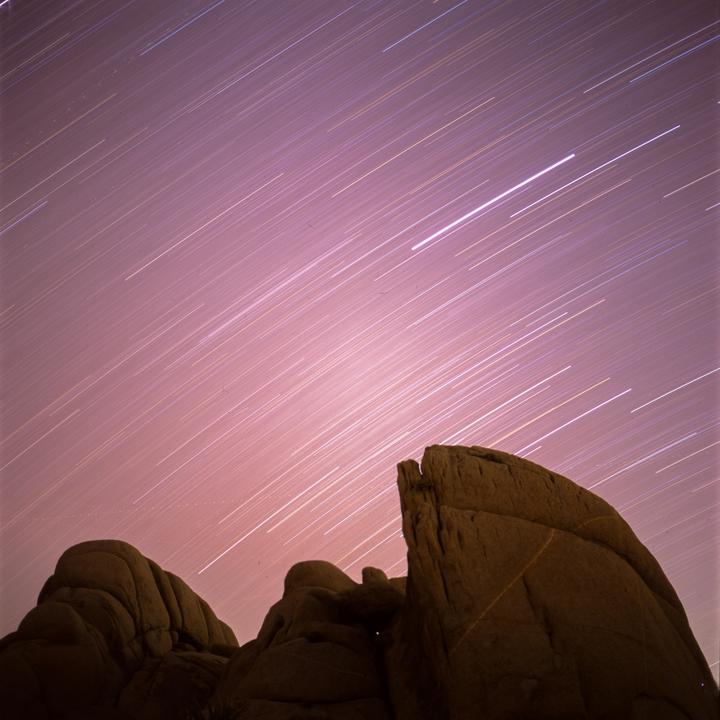 Star trails streak across a pink night sky above dark rock formations.