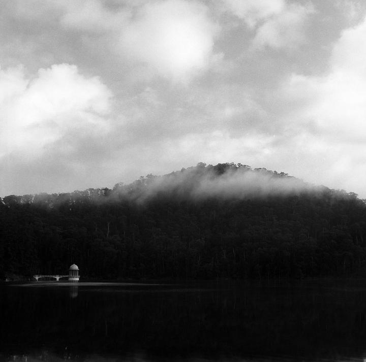 A small domed structure on a bridge extends into a lake with a forested, fog-covered hill in the background.