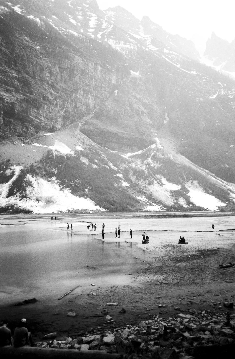 A group of people stand in a shallow body of water at the base of a large, snow-dusted mountain.