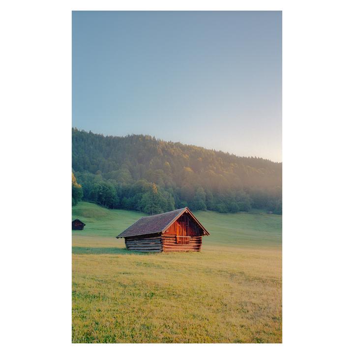 A wooden cabin sits in a grassy field with a forest and mountain in the background.