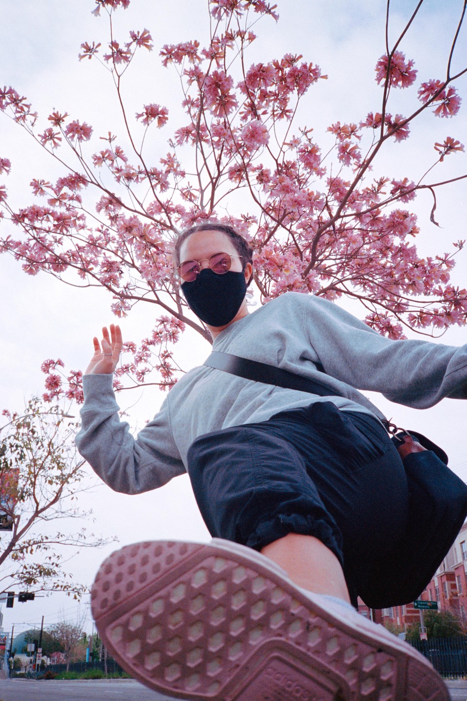 A person wearing a mask and sunglasses is captured from a low angle, with a blooming pink tree overhead and a street visible below.