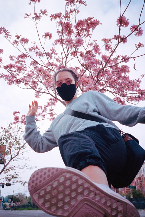 A person wearing a mask and sunglasses is captured from a low angle, with a blooming pink tree overhead and a street visible below.