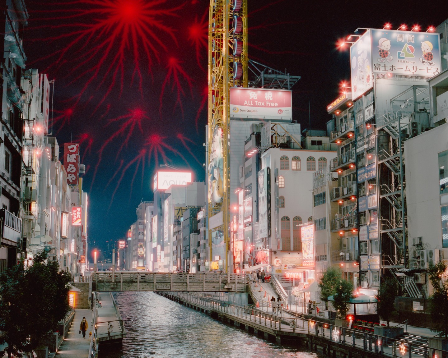 Red fireworks explode over a canal lined with buildings and neon signs at night.