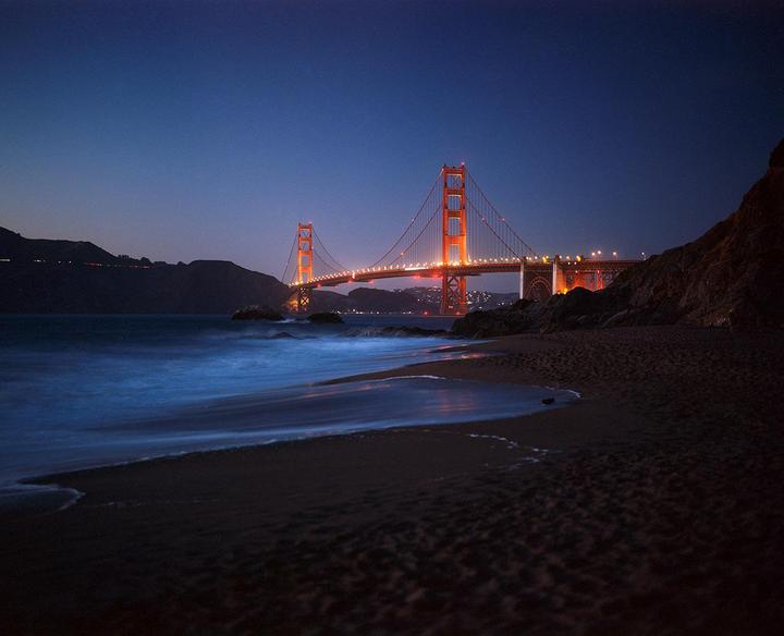 The Golden Gate Bridge is illuminated at night with a beach and ocean in the foreground.