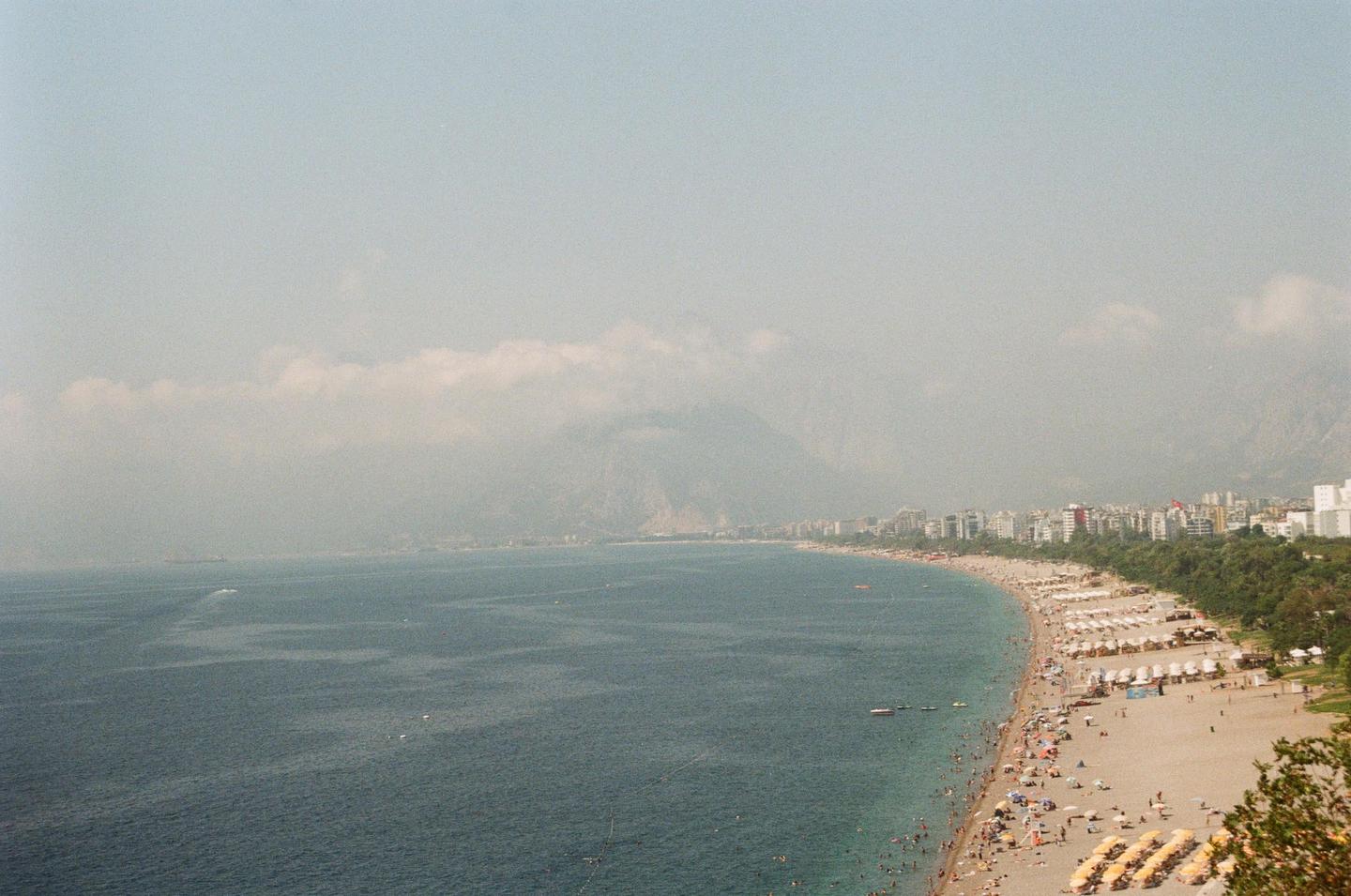A long beach with many people and umbrellas stretches along a coastline with mountains and buildings in the distance.