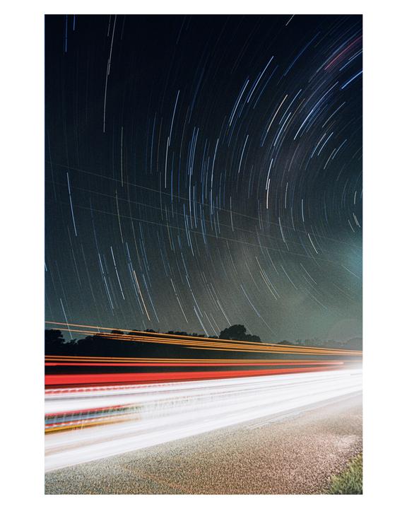 Star trails arc across the night sky above a road with streaks of light from passing vehicles.