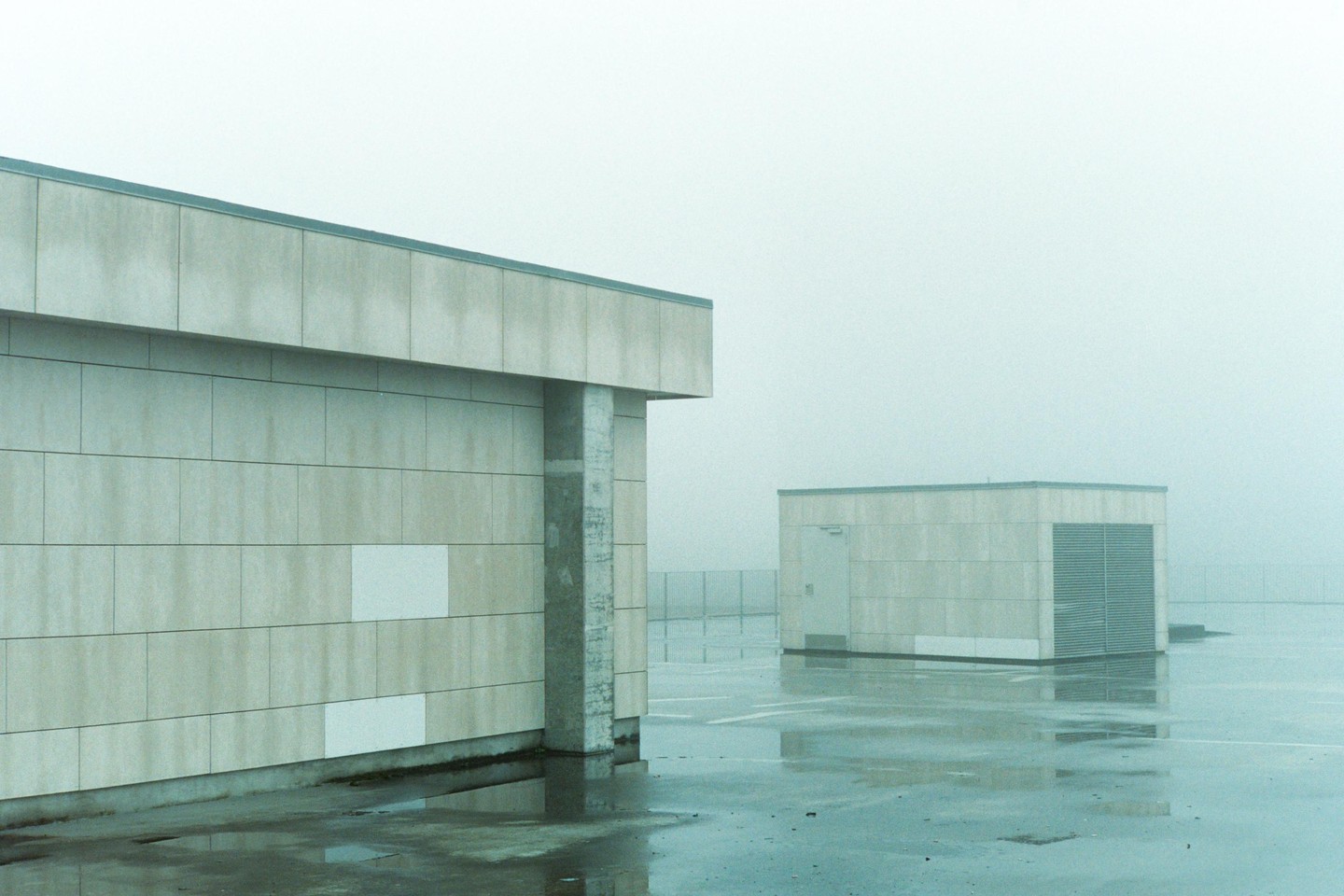 Two concrete buildings stand on a wet, paved surface under a hazy sky.