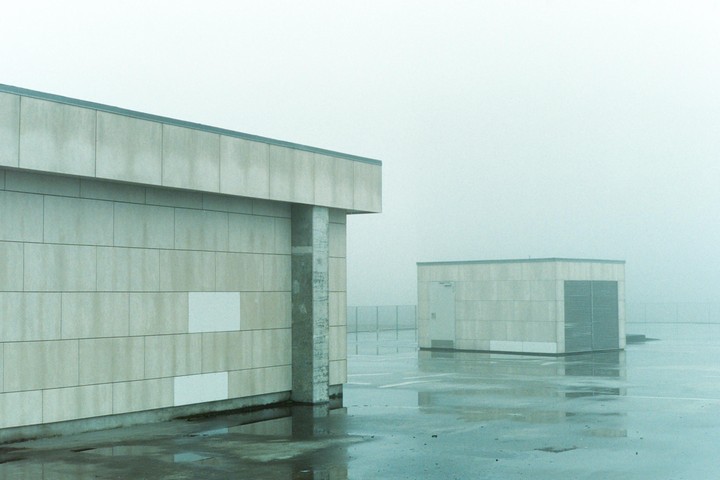 Two concrete buildings stand on a wet, paved surface under a hazy sky.