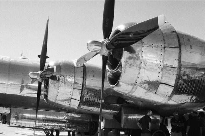 A large, shiny airplane with multiple propellers is parked on an airfield, with several men standing near its fuselage.
