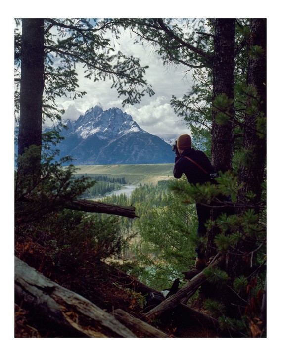 A man stands among trees, photographing a mountain range with a river and forest below.
