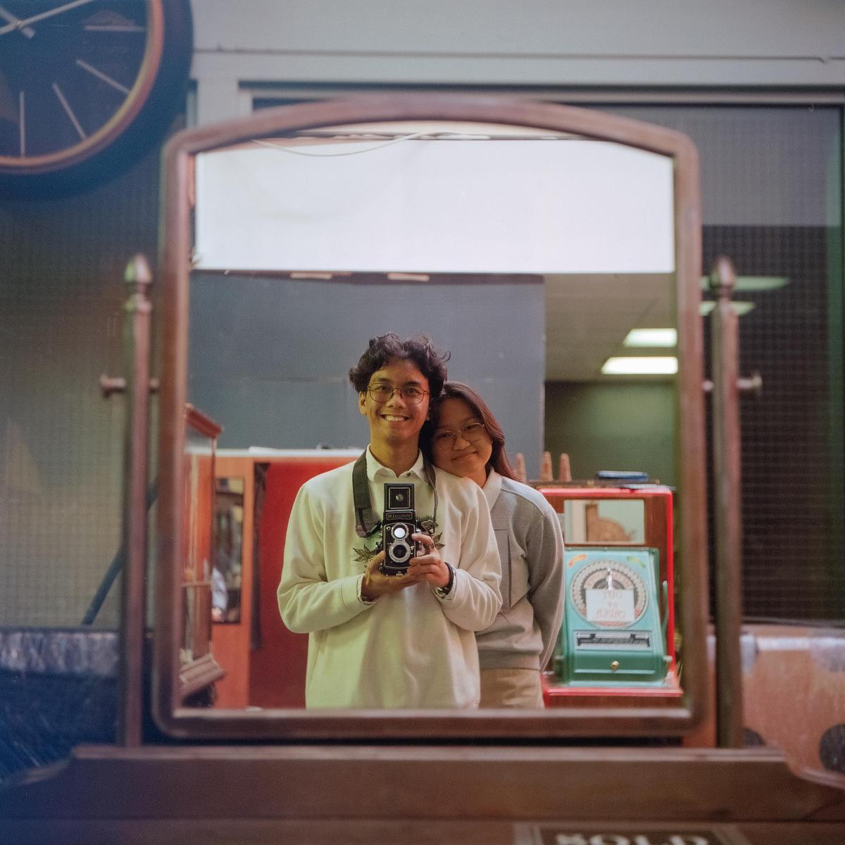 A man and a woman are reflected in a mirror, with the man holding a camera.