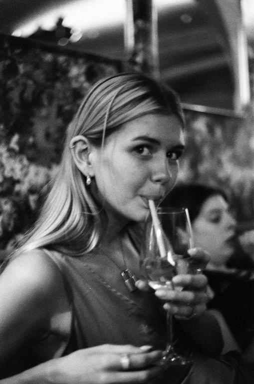 A woman with long hair looks at the viewer while drinking from a glass with a straw.