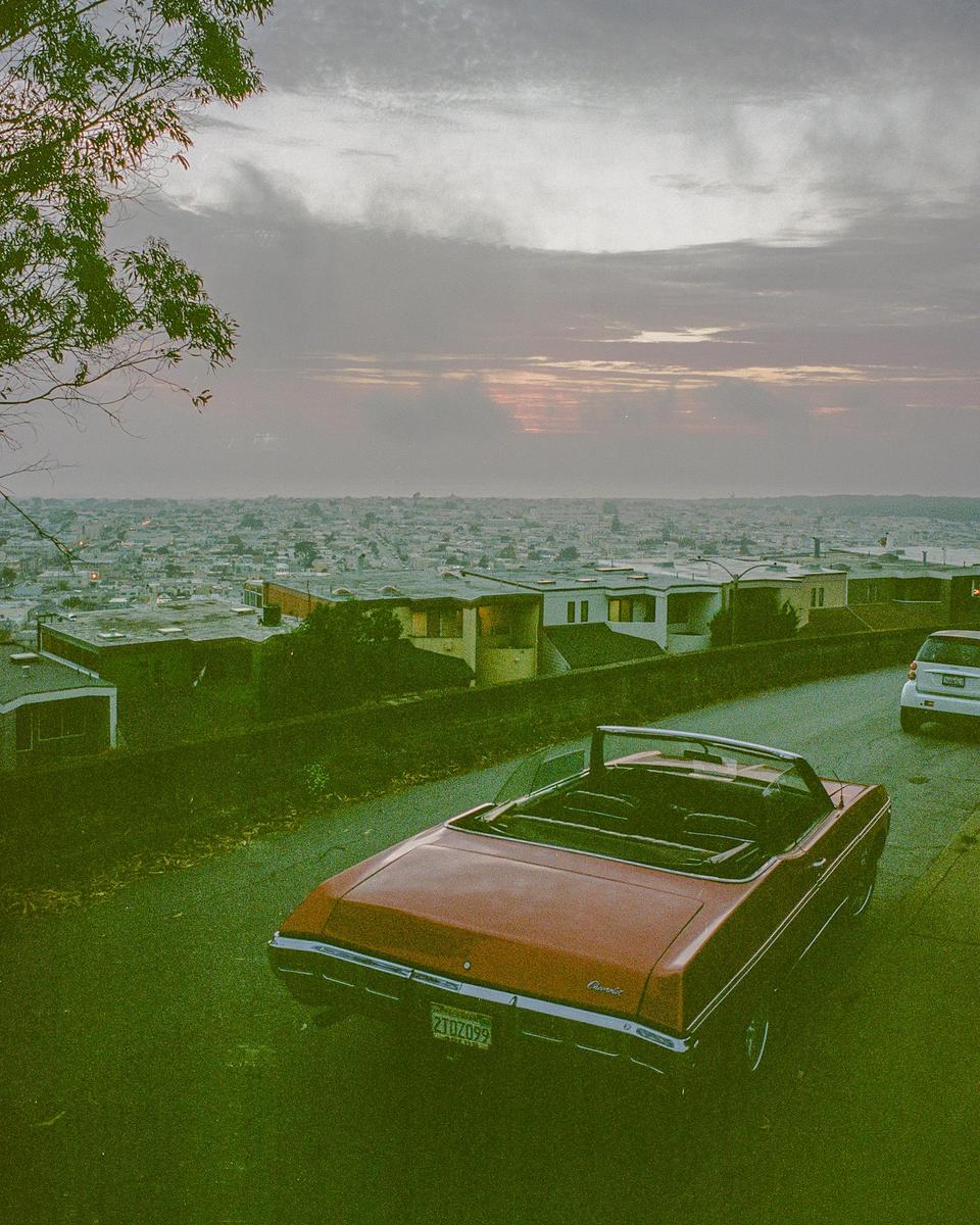 A red convertible is parked on a street overlooking a city at dusk.