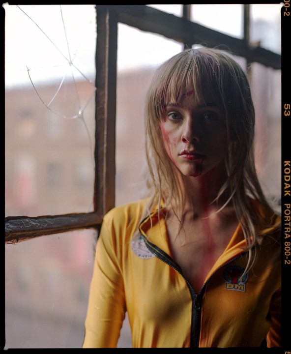 A woman with blonde hair and blood on her face wears a yellow jumpsuit and stands next to a broken window.