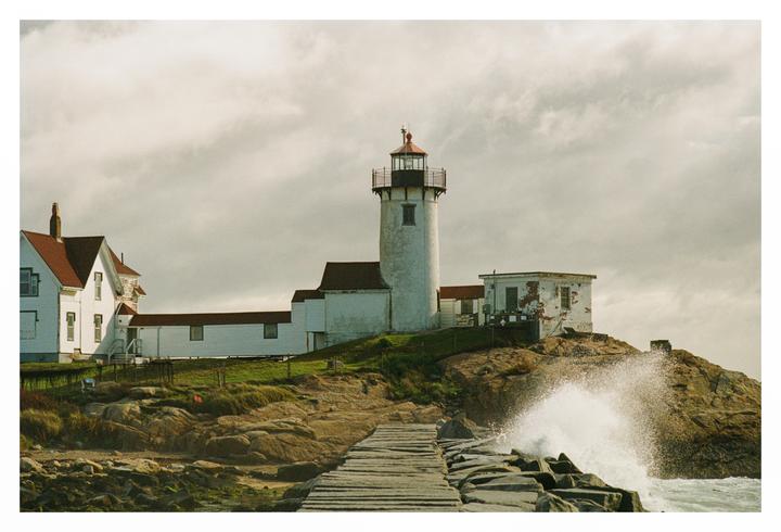 A white lighthouse and buildings stand on a rocky hill next to a wooden pier with a wave crashing against the rocks.