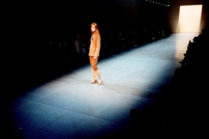 A woman in a brown suit walks down a runway in a fashion show.