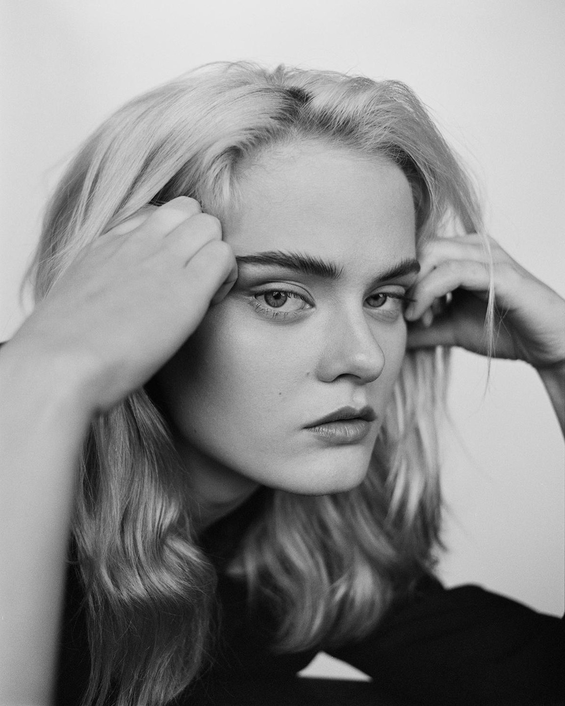 A woman with light hair holds her hands near her head and looks directly forward.
