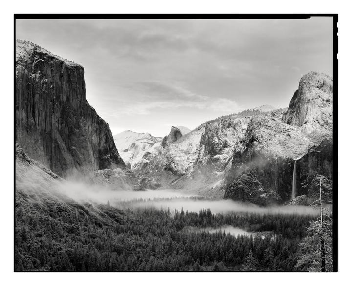 A valley with a forest and a waterfall is covered in fog and surrounded by mountains.