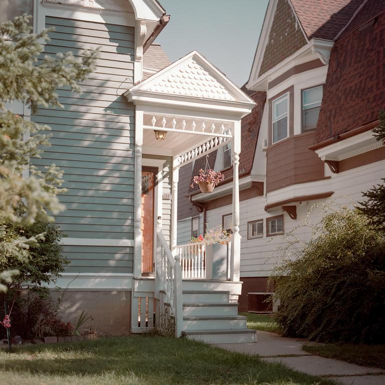 A white porch with steps leads to a house with light blue siding, next to a house with white siding and a brown roof.
