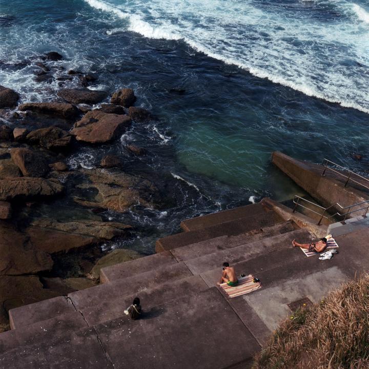 Three people are relaxing on concrete steps next to a rocky ocean shore with waves crashing.