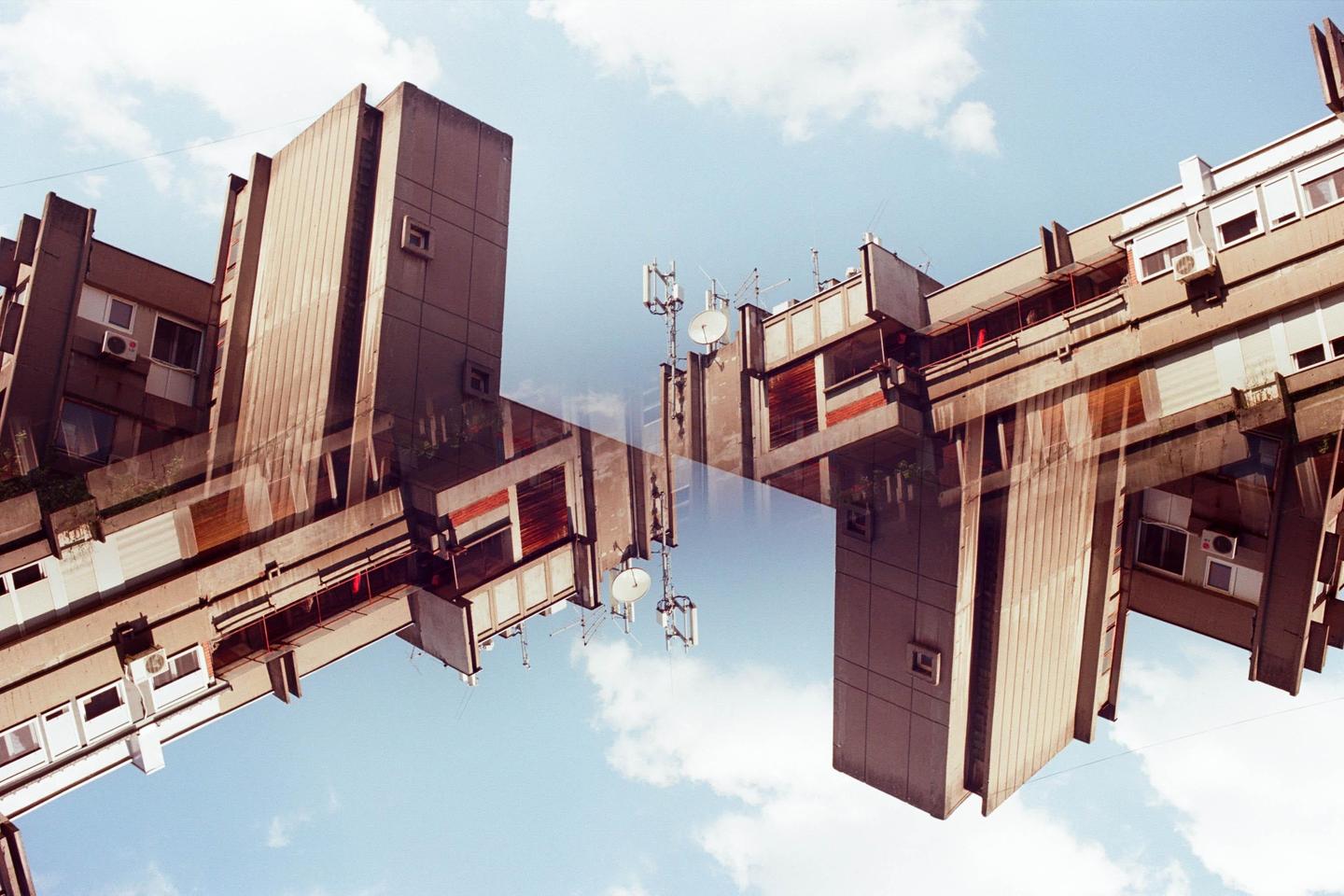A double exposure shows two concrete buildings against a blue sky with clouds.