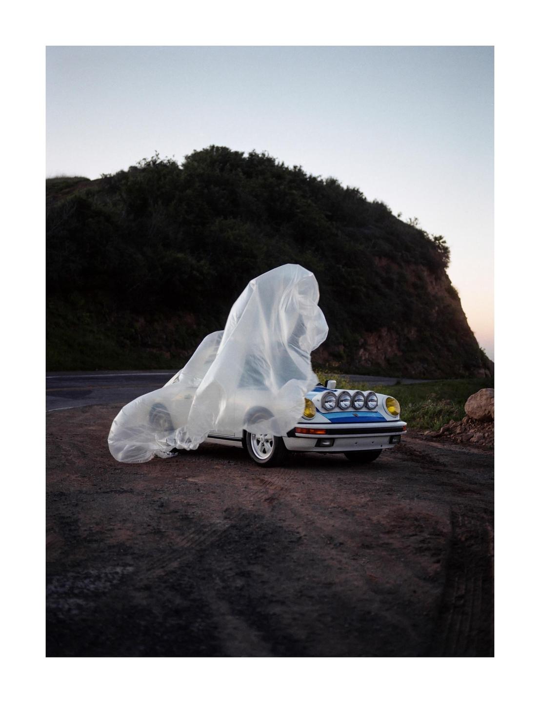 A white car with blue stripes and yellow headlights is parked on a dirt road next to a hill, partially covered by a translucent plastic sheet.